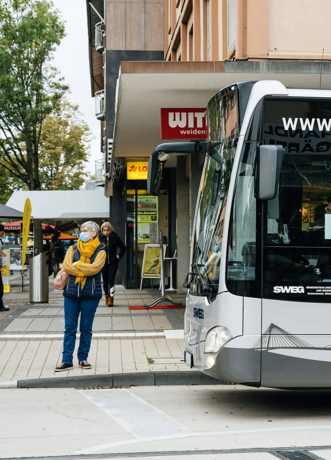 Eine Pendlerin steht an einer Bushaltestelle vor einem wartenden Bus.