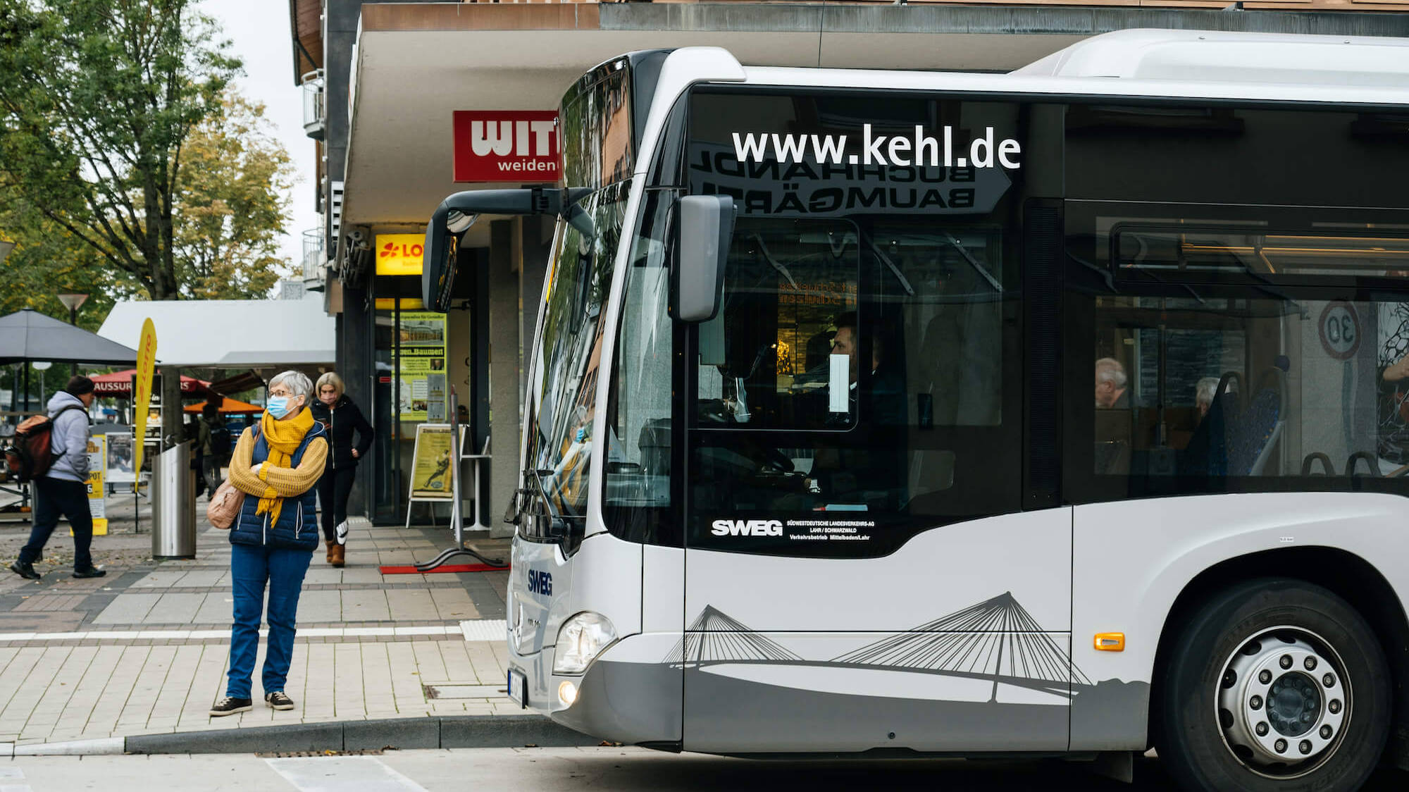 Eine Pendlerin steht an einer Bushaltestelle vor einem wartenden Bus.