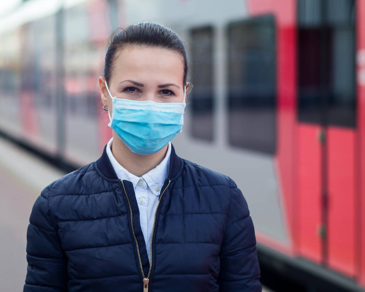 Eine Frau mit medizinischer Maske steht am Gleis vor einer Bahn