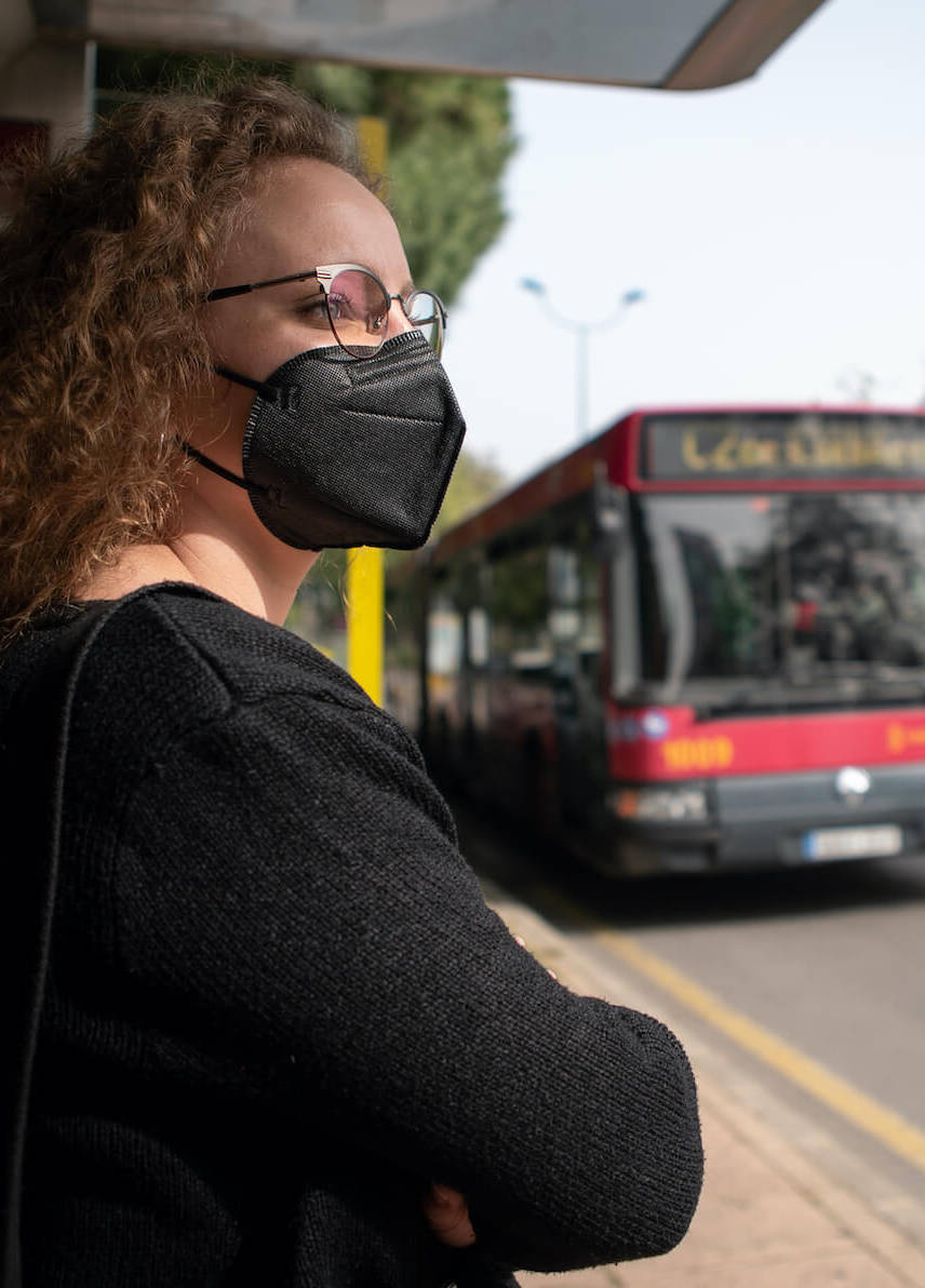 Frau mit FFP2-Maske steht an einer Bus-Haltestelle