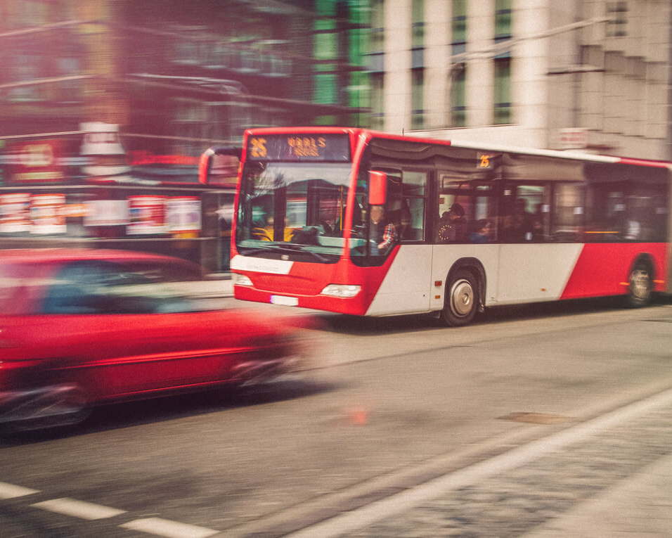 Ein roter Bus fährt an einem roten Auto in einer Großstadt vorbei. 